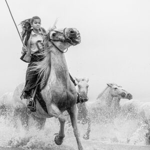 Photo en tirage limité en noir et blanc. Série chevaux de camargue de Michèle Gabet Photographie. jeune gardiane guidant son troupeau de juments au galop dans un marais